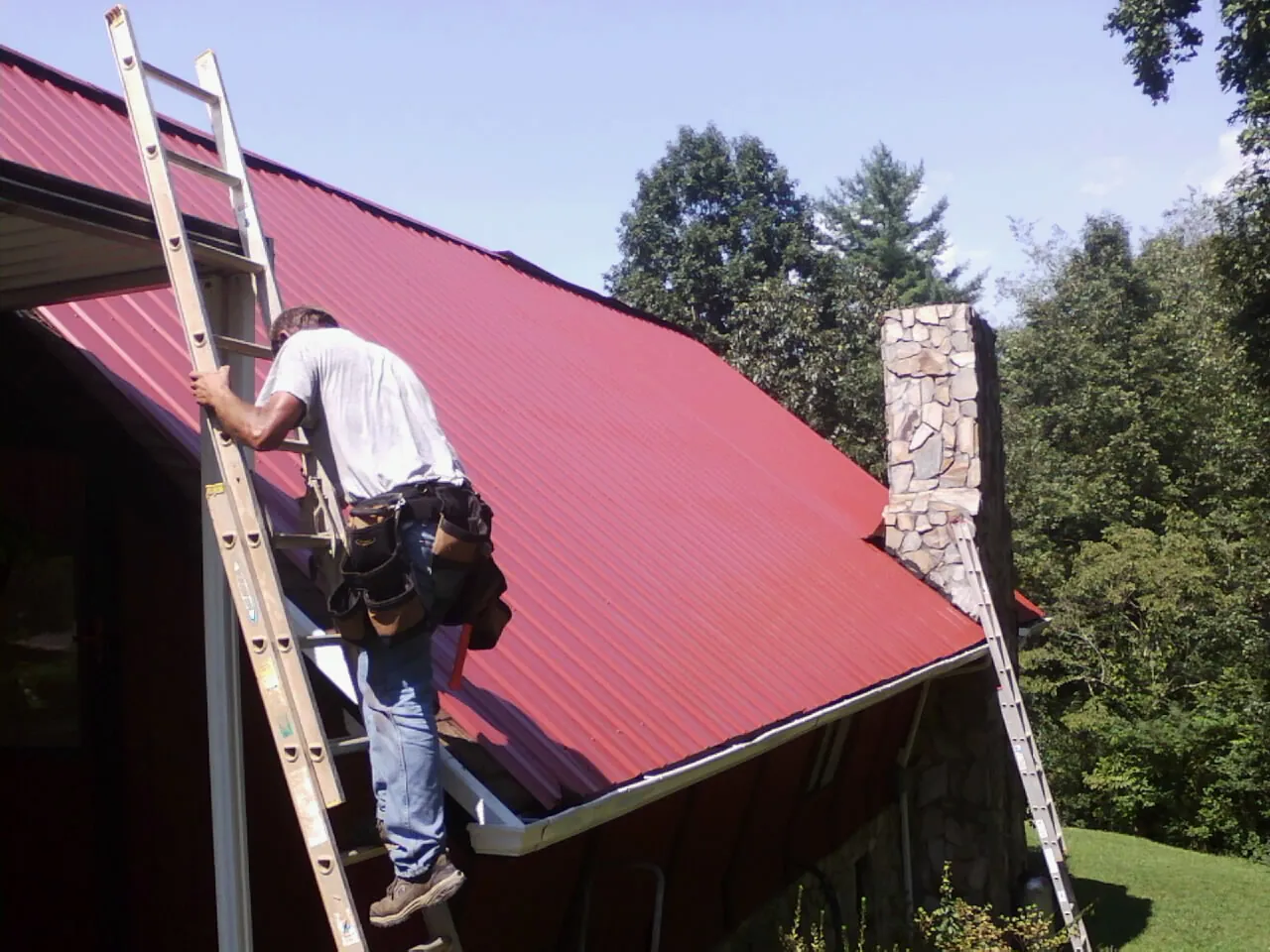 Roofer on a ladder beside a red metal roof and stone chimney — Soffit & Fascia Repair by Roofing Brothers in Clear Lake